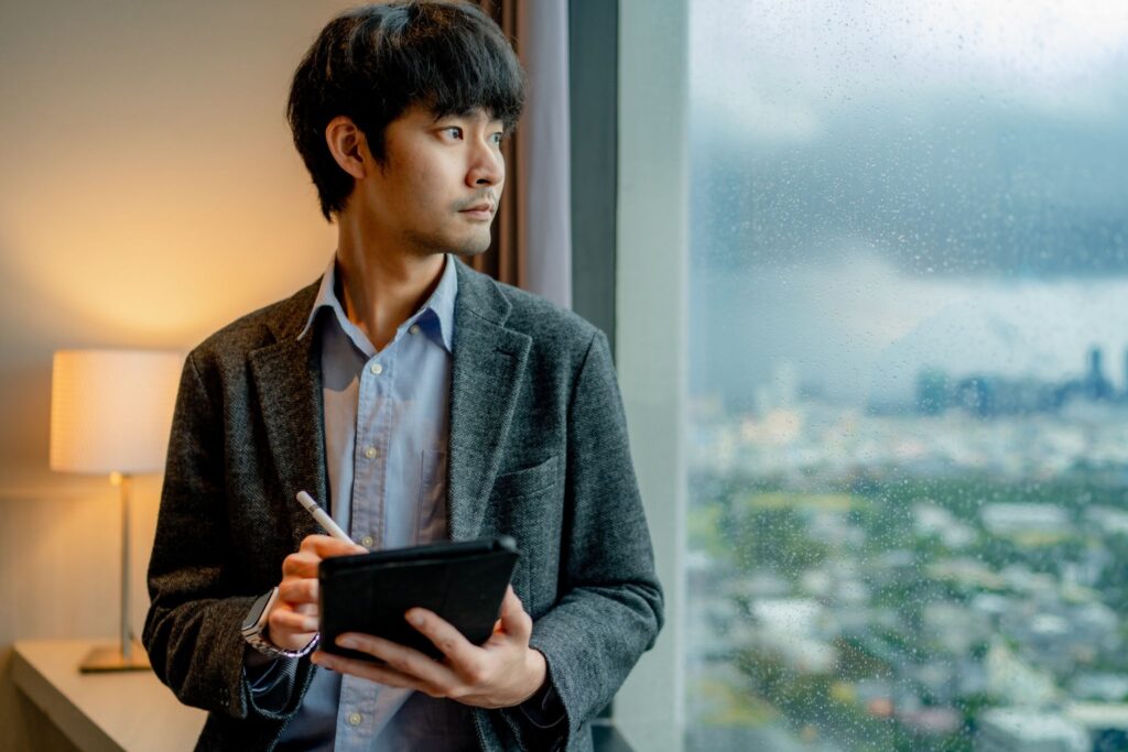 Young professional Asian man in a suit jacket, working on a tablet, looking out of a window.